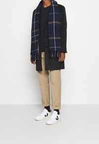 Man med beige uppvikta byxor, svart kappa, marinblå tröja, blå rutmönstrad scarf, svarta strumpor och vita sneakers som står mot en enfärgad bakgrund.