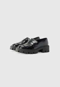 Black leather slip-on shoes with broad straps and metallic studs, featuring a chunky, textured black sole designed for stability.