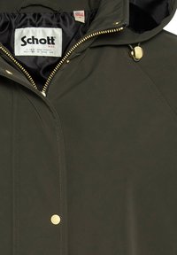 Giacca impermeabile verde oliva scuro con zip frontale, accessori dorati e una morbida fodera interna nera. Etichetta Schott visibile.