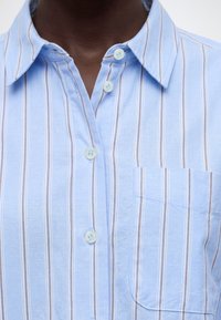 Gros plan d'une personne portant une chemise bleu clair avec des rayures beiges verticales, des boutons blancs et une poche poitrine sur le côté gauche.