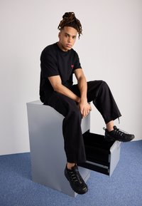 Schwarzes T-Shirt mit rotem Logo, schwarze Cargo-Hosen und schwarze Sneakers. Sitzt auf einer grauen Kommode mit einer offenen schwarzen Schublade. Minimalistischer Hintergrund.