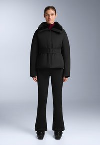 Crna jakna s naletom s lažnim krznom na ovratniku, remenom u struku i dugim rukavima, uparena s uskim crnim hlačama i sjajnim cipelama.