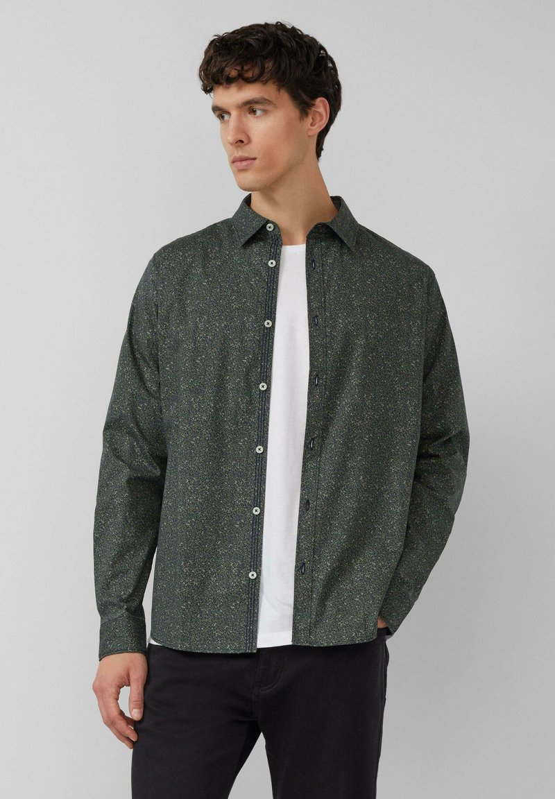 Chemise boutonnée vert foncé avec un motif moucheté, manches longues et col large, portée par-dessus un t-shirt blanc.