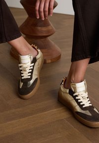 Brown and gold sneakers featuring a mix of suede and leather, with textured accents, thick laces, and a rubber sole. Logo on the heel.