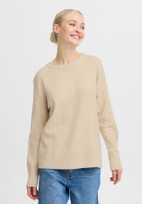 Maglione beige lavorato a maglia con scollatura tonda e polsini a costine. Il design presenta una vestibilità rilassata e una trama liscia, abbinato a jeans blu.