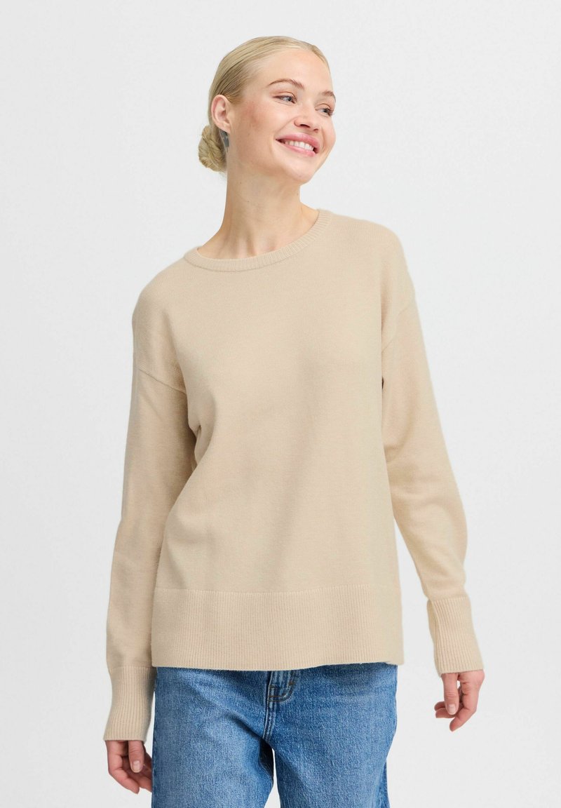 Maglione beige lavorato a maglia con scollatura tonda e polsini a costine. Il design presenta una vestibilità rilassata e una trama liscia, abbinato a jeans blu.