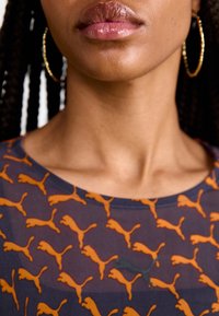 Mörkblå skjorta med ett heltäckande mönster av orange Puma-loggor, rund halsringning och korta ärmar. Stora ringörhängen kompletterar looken.