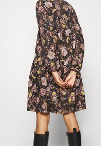 Schwarzes Kleid mit langen Ärmeln, Blumenmuster in Pink, Gelb und Weiß, geraffte Taille, ausgestellter Rock, getragen mit kniehohen schwarzen Stiefeln.