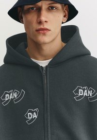 Szürke cipzáras kapucnis pulóver, több fehér hímzett logóval díszítve. Puha anyagból készült, hagyományos szabással és kapucnival.