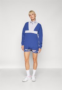 Person mit kurzen blonden Haaren, der einen blauen und hellgrauen sportlichen Pullover, passende Shorts, weiße Socken und weiße Sneaker trägt, steht vor einem weißen Hintergrund.