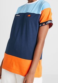 Tričko s barevnými bloky v odstínech světle modré, námořnické modré a oranžové. Vyrobeno z bavlny, s krátkými rukávy a logem na hrudi.
