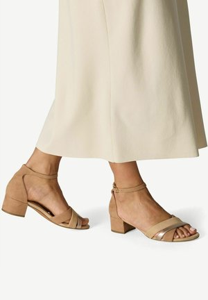 Pieds portant des sandales à talons blocs beiges avec des brides à la cheville, assorties à une jupe midi ou une robe de couleur crème.