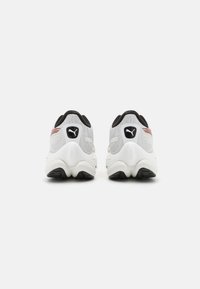 Zapatillas deportivas blancas con una parte superior de malla texturizada, acentos negros y una suela gruesa con un dibujado de patrón. Logo visible en el talón.