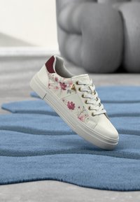 Baskets blanches avec motif floral rose et détail de talon bordeaux reposant sur un tapis bleu texturé dans un cadre intérieur moderne.