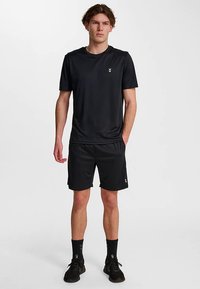 Svart sport-t-shirt och shorts i lättviktsmaterial med en liten logotyp. Modellen står med händerna i fickorna.