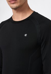 Nahaufnahme einer Person, die ein schwarzes, langärmliges Sportshirt mit einem kleinen weißen Champion-Logo auf der Brust trägt.