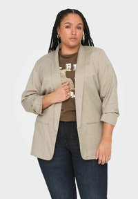 Helebeežž värvi kohandatud bleiser, millel on lõikega kraed, kolmandiku pikkuses varrukad ja ees taskud, kihistatuna pruunist graafilisest T-särgist ja tumedatest teksadest.