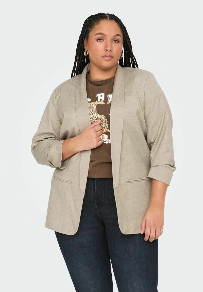 Helebeežž värvi kohandatud bleiser, millel on lõikega kraed, kolmandiku pikkuses varrukad ja ees taskud, kihistatuna pruunist graafilisest T-särgist ja tumedatest teksadest.