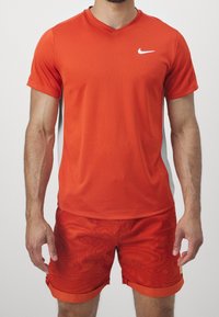 Červené sportovní tričko s výstřihem do V a bílým logem, doplněné červenými šortkami s vzorem a manžetovým lemem. Oba kusy jsou lehké a prodyšné.