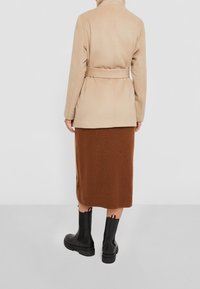 Beige mantel van een wolmix met een tailleband, gecombineerd met een bruine geribbelde gebreide rok en zwarte enkellaarzen met kleurrijke accenten.