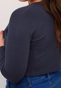 Marinefarbener, verkürzter Cardigan mit langen Ärmeln, strukturierter Strickware, schmaler Rippung entlang der Kanten und taillierter Passform. Über Denim getragen.