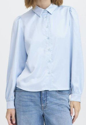 Blouse bleu clair à boutons avec manches longues bouffantes portée avec un jean en denim bleu sur une personne sur fond blanc.