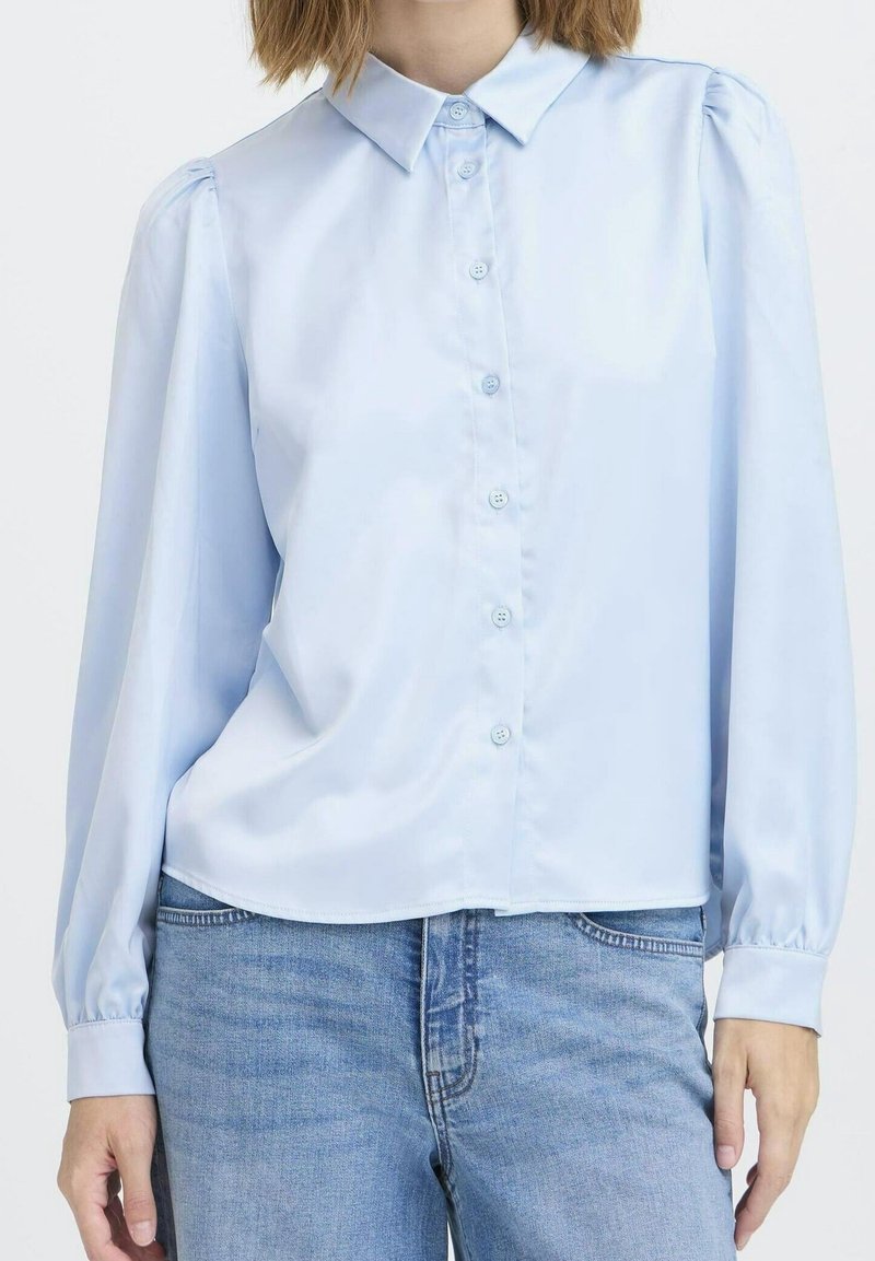 Blouse bleu clair à boutons avec manches longues bouffantes portée avec un jean en denim bleu sur une personne sur fond blanc.