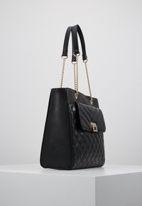 Czarna pikowana skórzana torebka na ramię ze złotymi łańcuszkowymi paskami i przednią kieszenią z klapą, stojąca na białym piedestale na jednolitym szarym tle.