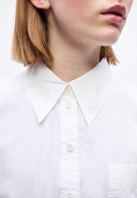 Γυναίκα με κοντά ανοιχτό καστανά μαλλιά που φοράει καθαρό λευκό πουκάμισο με κουμπιά και μυτερό γιακά, το κεφάλι γερμένο ελαφρώς στο πλάι.