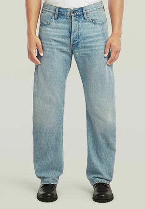 Lichtblauwe jeans met rechte pijpen gedragen met zwarte laarzen, gestyled met handen ontspannen langs het lichaam.
