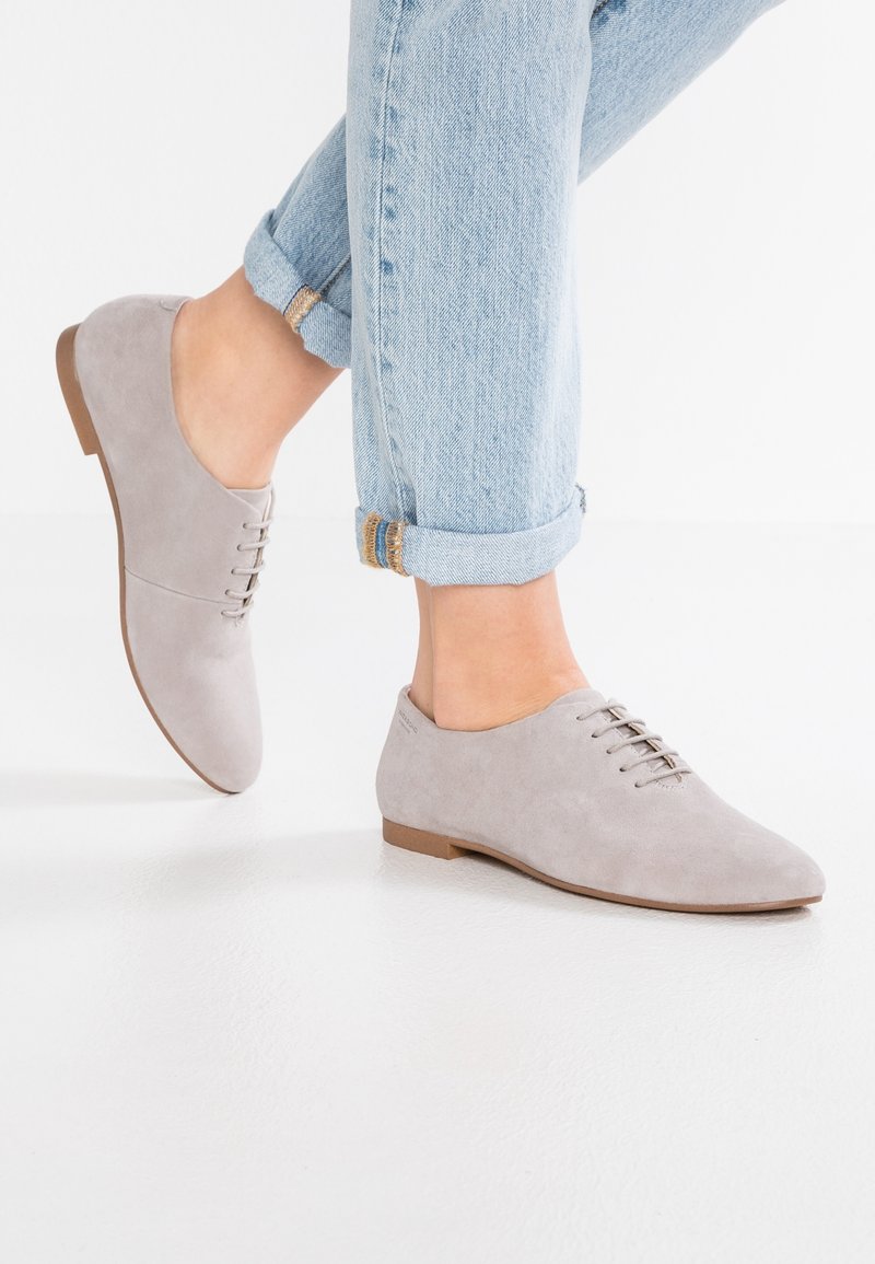 Chaussures Oxford en daim gris clair à lacets portées avec un jean bleu clair retroussé sur un fond blanc uni.