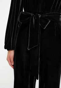 Czarny welurowy kombinezon z długimi rękawami, z dopasowaną talią i dużą związaną kokardą jako akcentem. Materiał ma miękką, błyszczącą teksturę.