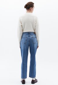 Krämfärgad tröja, blå högmidjade jeans och bruna loafers. Jeansen har rak passform och klassiska bakfickor. Lätt textur överlag.