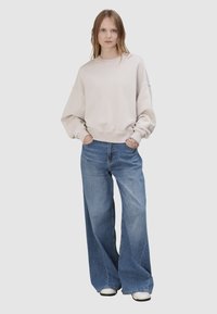 Kobieta w oversize'owej beżowej bluzie i szerokich niebieskich dżinsach, z rękami w kieszeniach, stojąca na tle jednolitego jasnego tła.