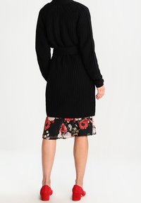 Knitted black long cardigan with a belted waist, paired with a floral pleated skirt. Red block-heeled shoes complete the outfit.