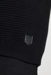 Černá žebrovaná pletenina s malou černou koženou nášivkou přišitou blízko lemu přes černou džínovou látku.