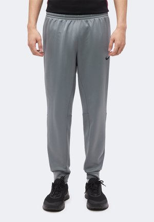 Homme portant un pantalon de jogging gris Nike et des baskets noires, debout devant un fond blanc uni.