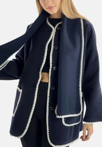 Chaqueta azul marino de tela texturizada con ribete blanco con borde ondulado, que presenta grandes botones, un bolsillo y una bufanda a juego colgando del cuello.