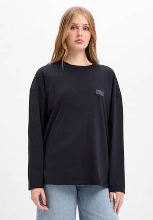 Jonge vrouw met lang blond haar die een zwart shirt met lange mouwen en lichtblauwe spijkerbroek draagt voor een witte achtergrond.