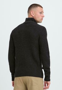 Mørkegrå strikket sweater med en høj krave, der har et struktureret mønster og ribbede manchetter, parret med beige bukser.