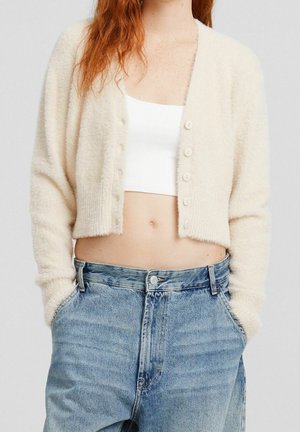 Femme portant un cardigan crémeux et duveteux sur un crop top blanc et un jean bleu taille haute, les mains dans les poches, sur fond uni.