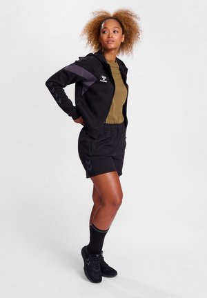 Schwarze Zip-up-Hoodie mit grauen Akzenten, olivgrünes T-Shirt, schwarze Shorts und schwarze Sportschuhe. Umgeschlagene schwarze Socken und lockiges Haar.
