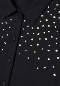 Camicetta nera con colletto e bottoni, decorata con piccoli strass trasparenti sparsi in varie forme sulla parte anteriore del tessuto.
