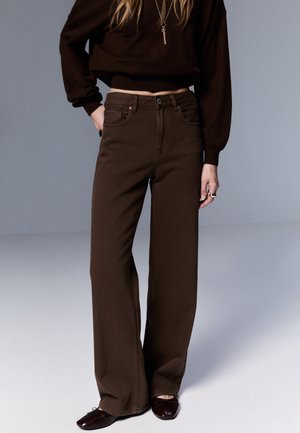 Smeđe široke traperice od trapera; imaju visok struk, prednje džepove i glatku teksturu, uparene s tamnim džemperom i ravnim cipelama.