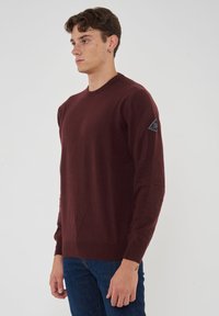 Maglione lavorato a maglia bordeaux con maniche lunghe, scollo rotondo e una toppa a logo triangolare sulla spalla sinistra. Texture liscia, design aderente.