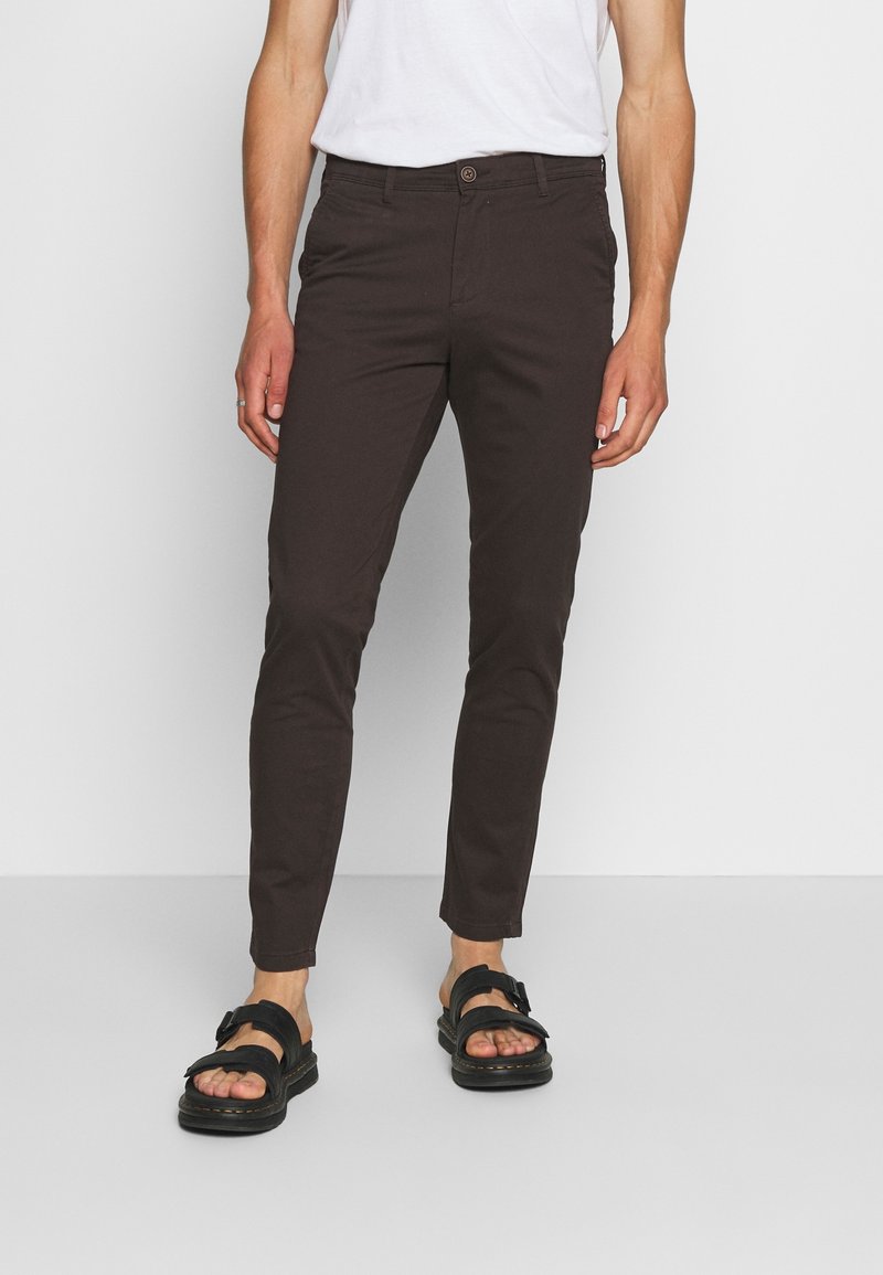 Ruskeat, kapealahkeiset puuvillahousut, joissa on sileä pinta ja yksittäinen napin suljenta, yhdistetty mustiin sandaaleihin.