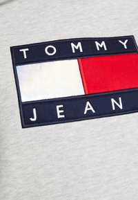 Tejido gris con un parche rectangular azul marino que lleva bordado en blanco el texto "TOMMY" por encima de un diseño dividido de paneles rojos y reflectantes en blanco.
