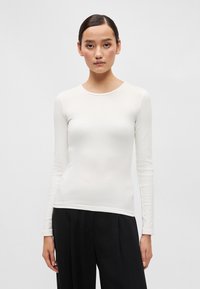 Fehér hosszú ujjú ing kerek nyakkivágással. Feszes fazon sima anyagból, magas derekú fekete nadrággal párosítva.