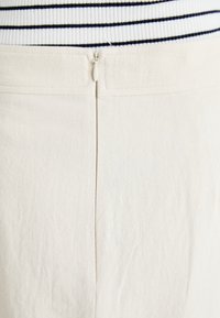 Pantaloni beige con texture liscia, dotati di una cerniera nascosta sul retro. Sezione superiore mostrata con maglione a righe bianche e nere.