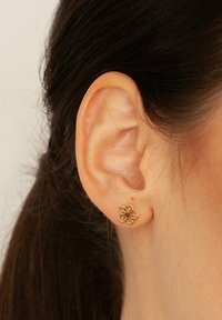 Złoty kolczyk w kształcie kwiatu z cienkim konturem, posiadający pięć płatków i mały detal w centrum. Noszony w przekutym uchu.